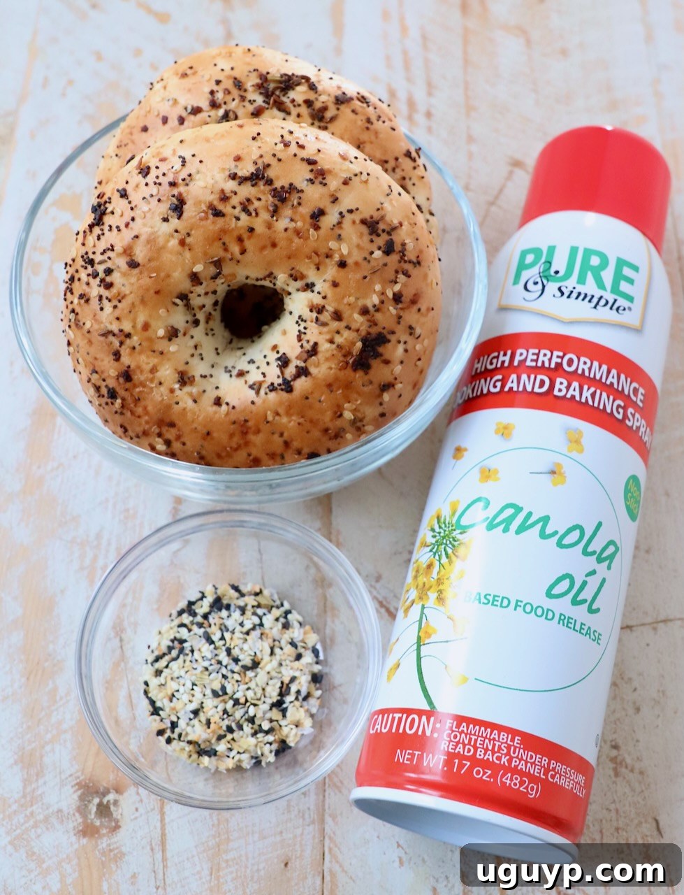 Ingredients for making bagel chips laid out on a white wooden board, including bagels, cooking spray, and everything bagel seasoning.
