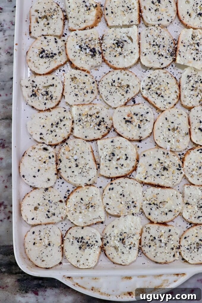 Thinly sliced bagel chips seasoned with everything bagel seasoning on a baking sheet.