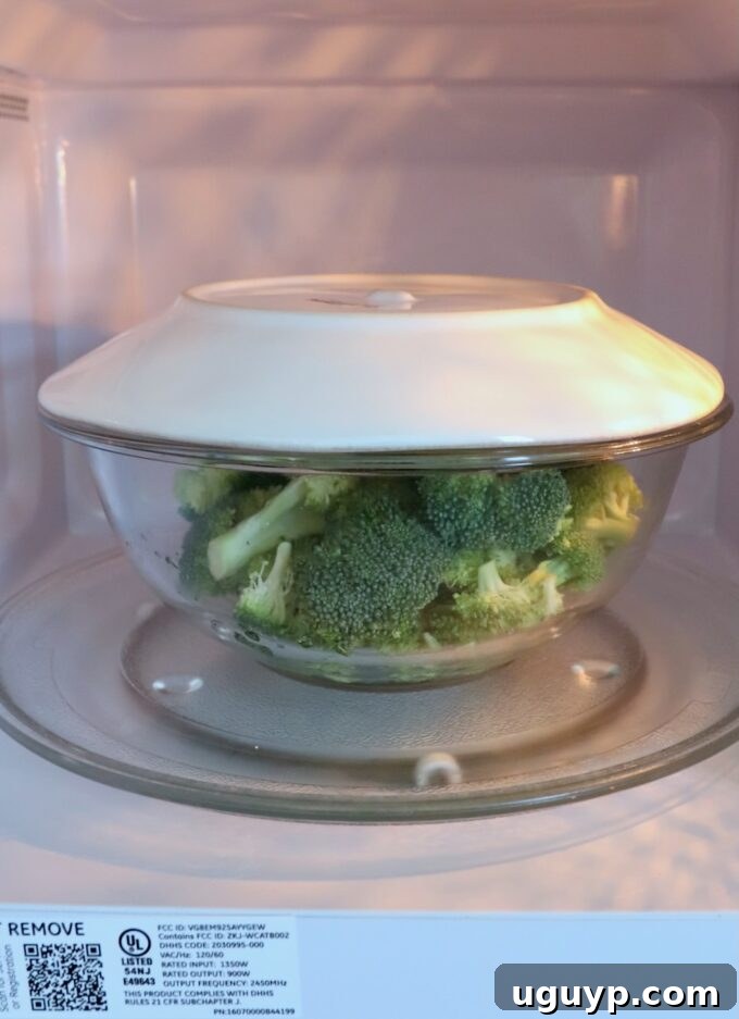 Broccoli florets in a glass bowl, ready for microwave steaming with a plate on top.
