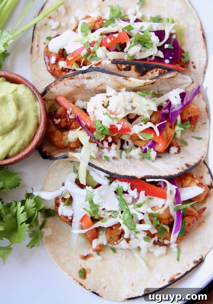 Overhead shot of three perfectly assembled grilled shrimp tacos, complemented by a side of creamy avocado salsa, on a white plate.