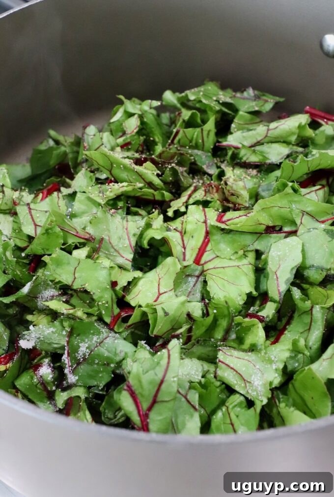 Fresh beet greens added to a large pan with sautéed aromatics, beginning to wilt.