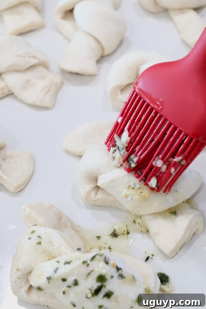 A pastry brush applies seasoned butter to uncooked garlic knots on a baking sheet, preparing them for the oven.