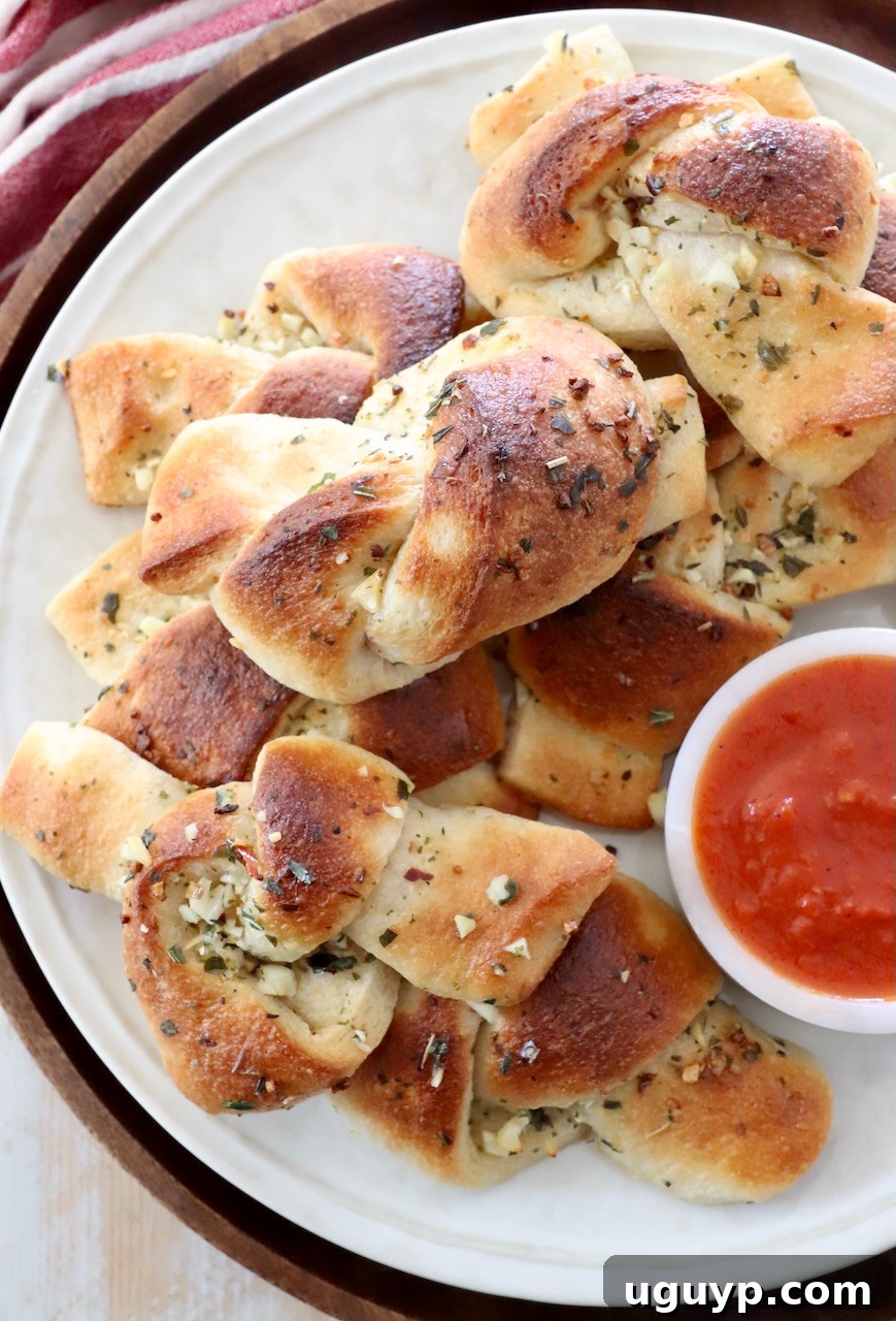 A stack of golden-brown garlic knots on a plate, with a small bowl of marinara sauce in the background.