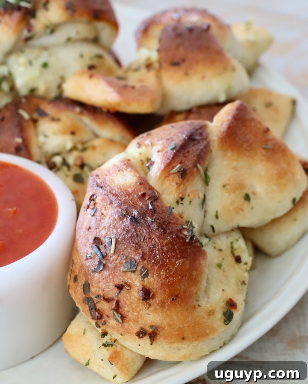 A serving of garlic knots on a white plate, accompanied by a small container of marinara dipping sauce.