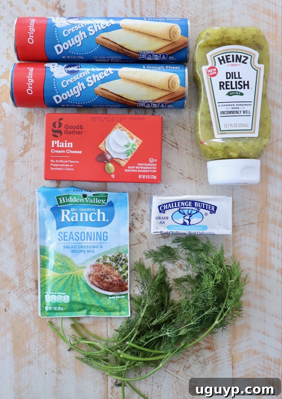 Ingredients for creamy pickle roll ups laid out on a white wooden board, including cream cheese, ranch seasoning, dill relish, crescent rolls, butter, and fresh dill.