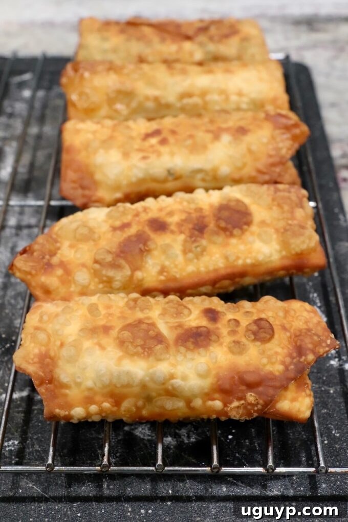 Freshly fried avocado egg rolls resting on a wire rack set over a cutting board, allowing excess oil to drip away and maintaining crispness.