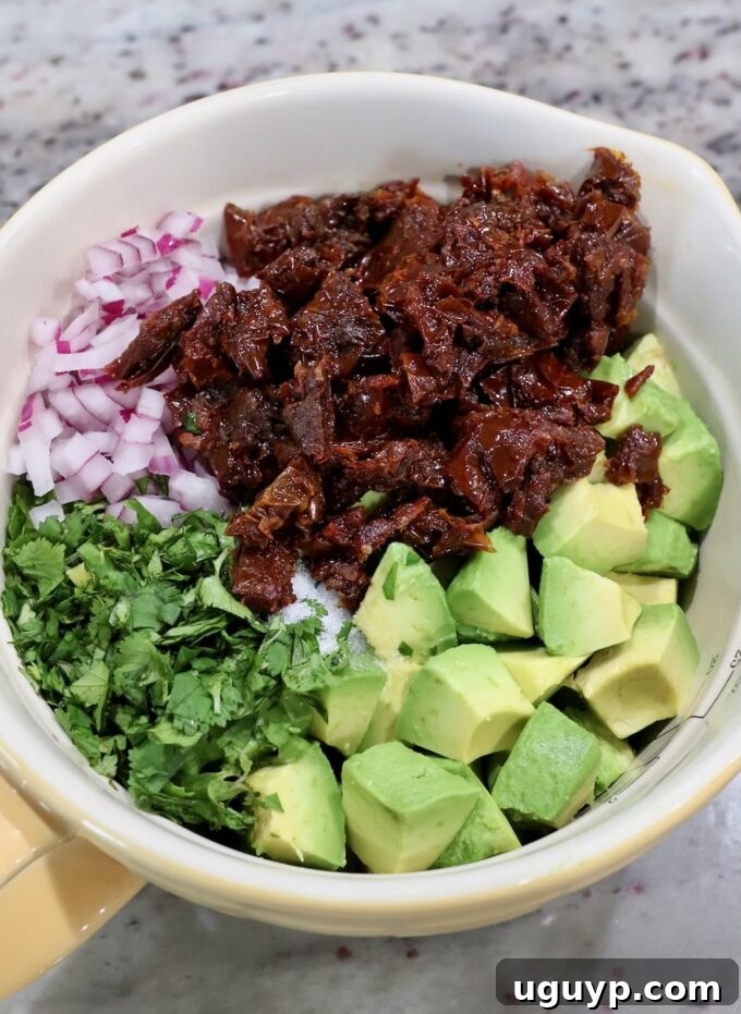 A bowl filled with diced avocado, finely chopped sun-dried tomatoes, minced red onion, and fresh cilantro, ready to be mixed for the egg roll filling.