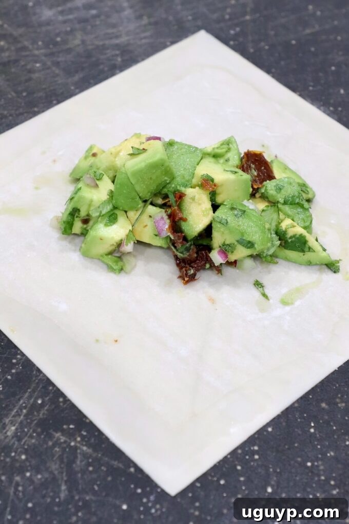An egg roll wrapper laid out diagonally on a cutting board with a spoonful of avocado mixture placed in the center, ready for folding.