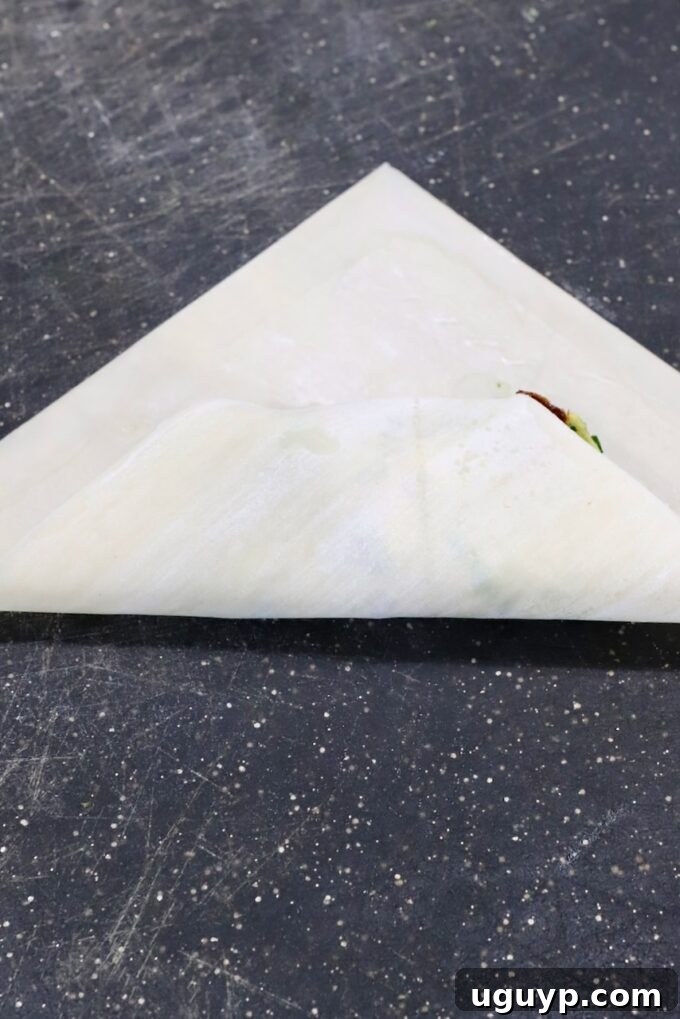 The first fold of the egg roll wrapper, with the bottom corner pulled up over the avocado filling and tucked underneath, creating a neat parcel.