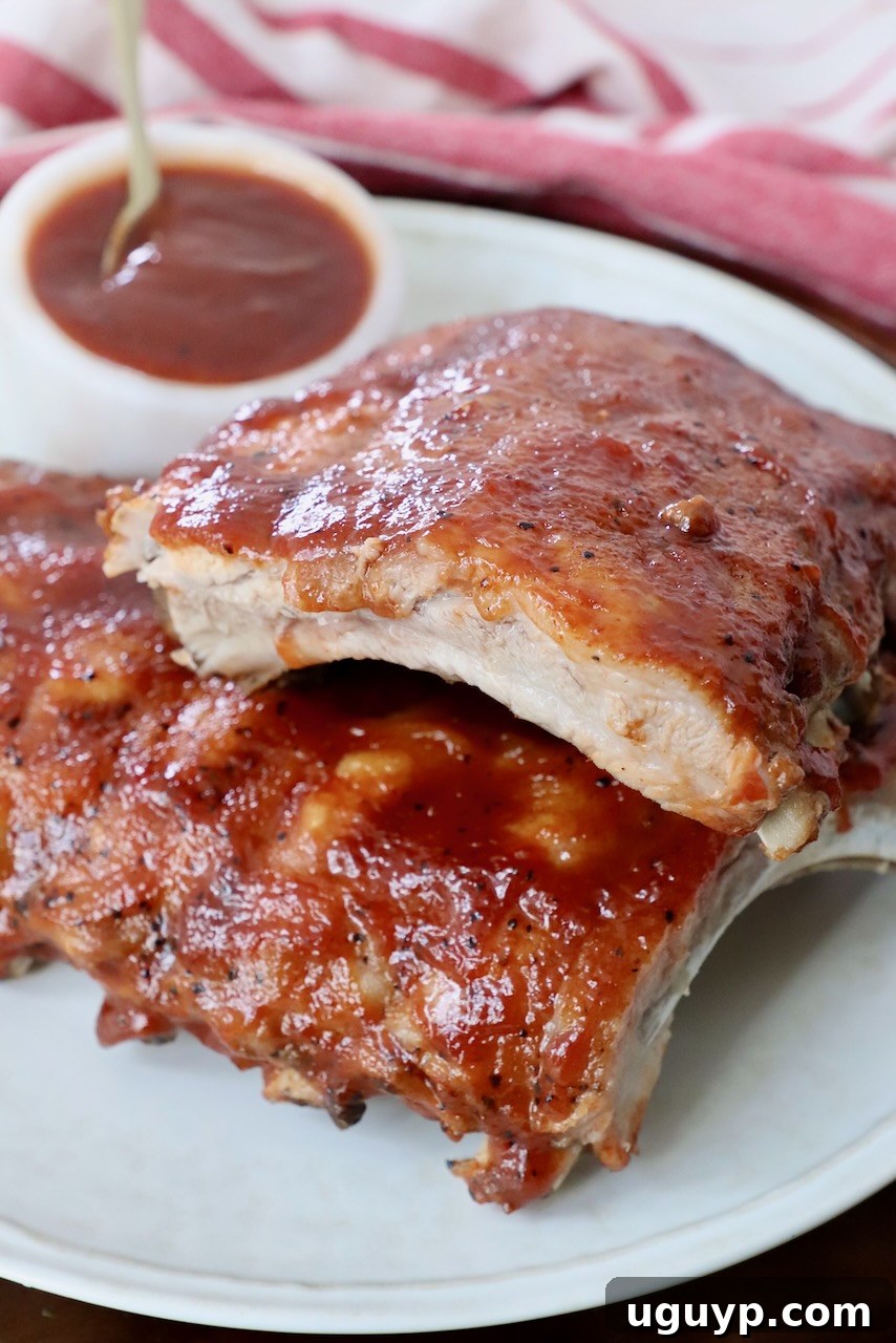 Cooked baby back ribs stacked on a plate with a small bowl of BBQ sauce on the side, ready to be served.