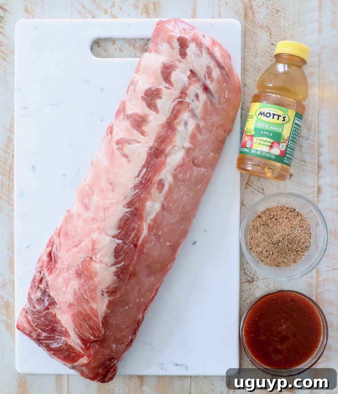 Ingredients for Instant Pot Ribs laid out on a white wooden board, including a rack of ribs, apple juice, dry rub, and BBQ sauce.