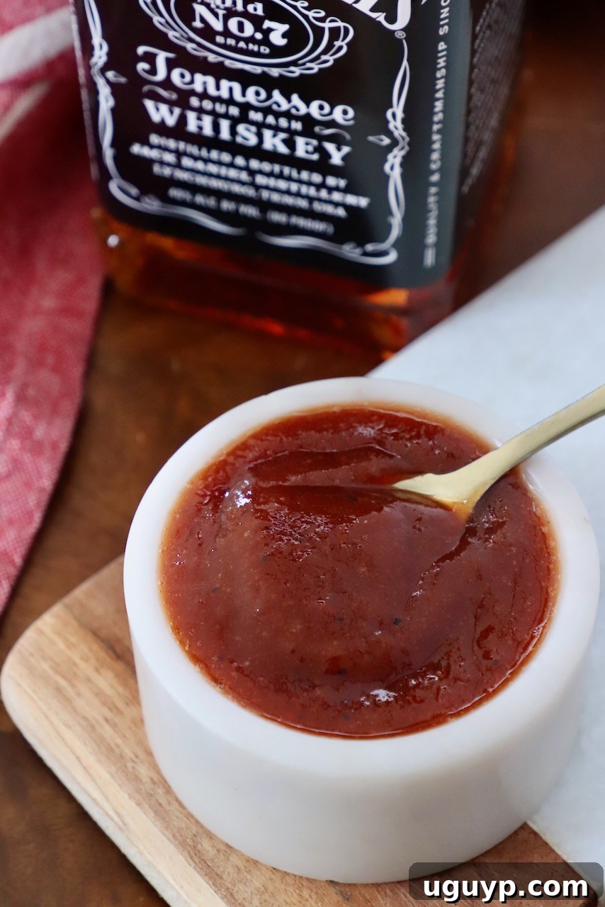 Homemade Jack Daniel's BBQ sauce in a small ceramic bowl with a rustic spoon, a classic bottle of Jack Daniel's Tennessee Whiskey subtly visible in the background, hinting at the sauce's key ingredient. The rich, dark glaze of the sauce is prominent.