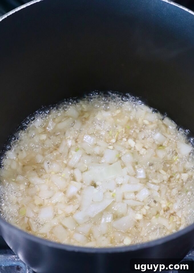 Diced yellow onions simmering in a saucepan with Jack Daniel's whiskey on a stovetop burner. The liquid is reducing and the onions are becoming translucent, beginning the infusion process for the BBQ sauce's base.