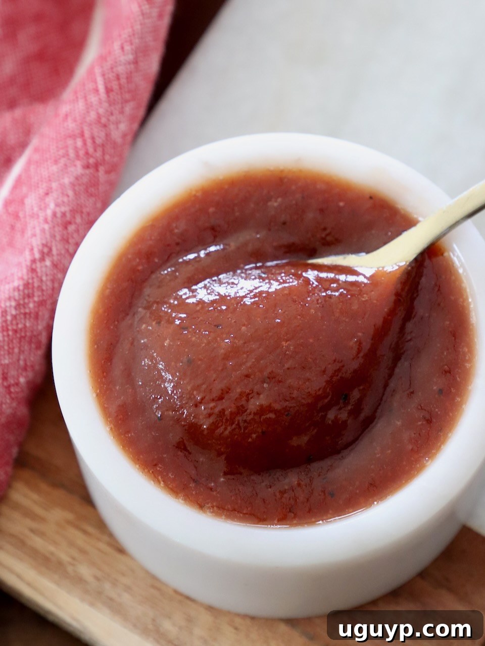 A small, elegant ceramic bowl filled with glistening, rich Jack Daniel's BBQ sauce, with a tiny spoon resting on the side, ready to be sampled or served. The sauce has a smooth, thick consistency and a deep, inviting color.