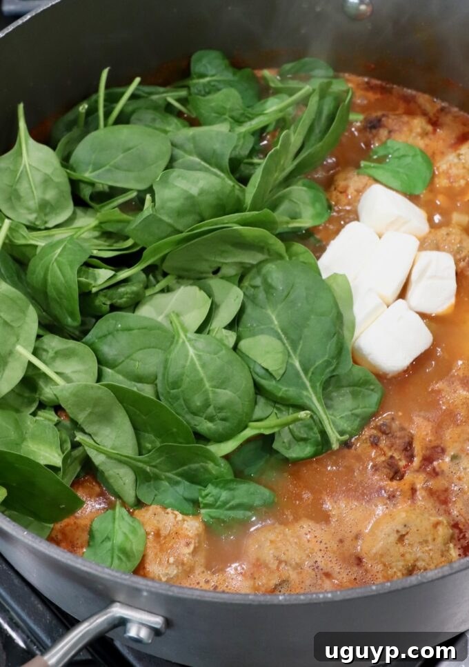 Fresh spinach leaves and cubes of cream cheese being added to the hot buffalo chicken meatball soup.