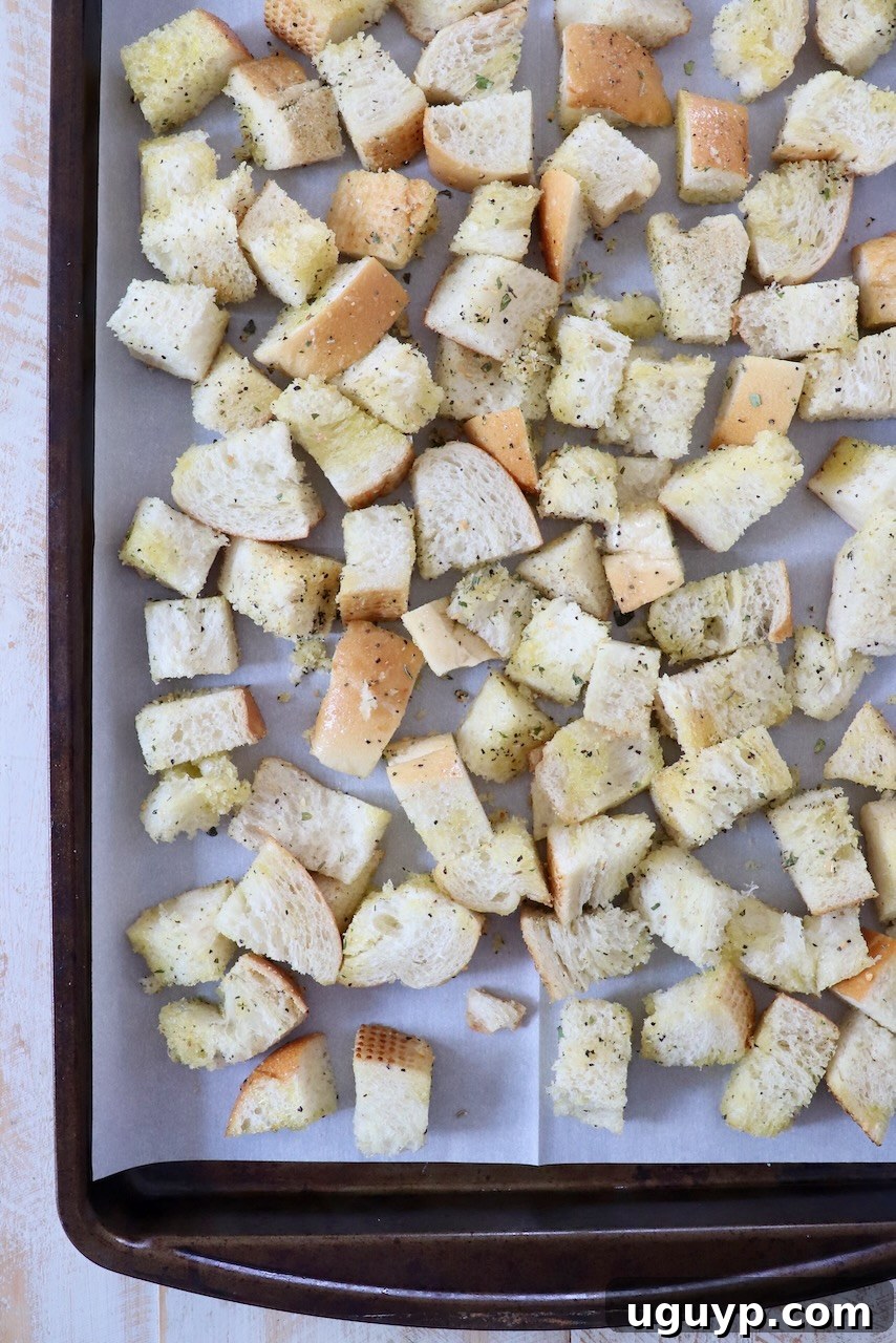 Cubed bread seasoned with herbs and spices, spread evenly on a parchment-lined baking sheet before baking.