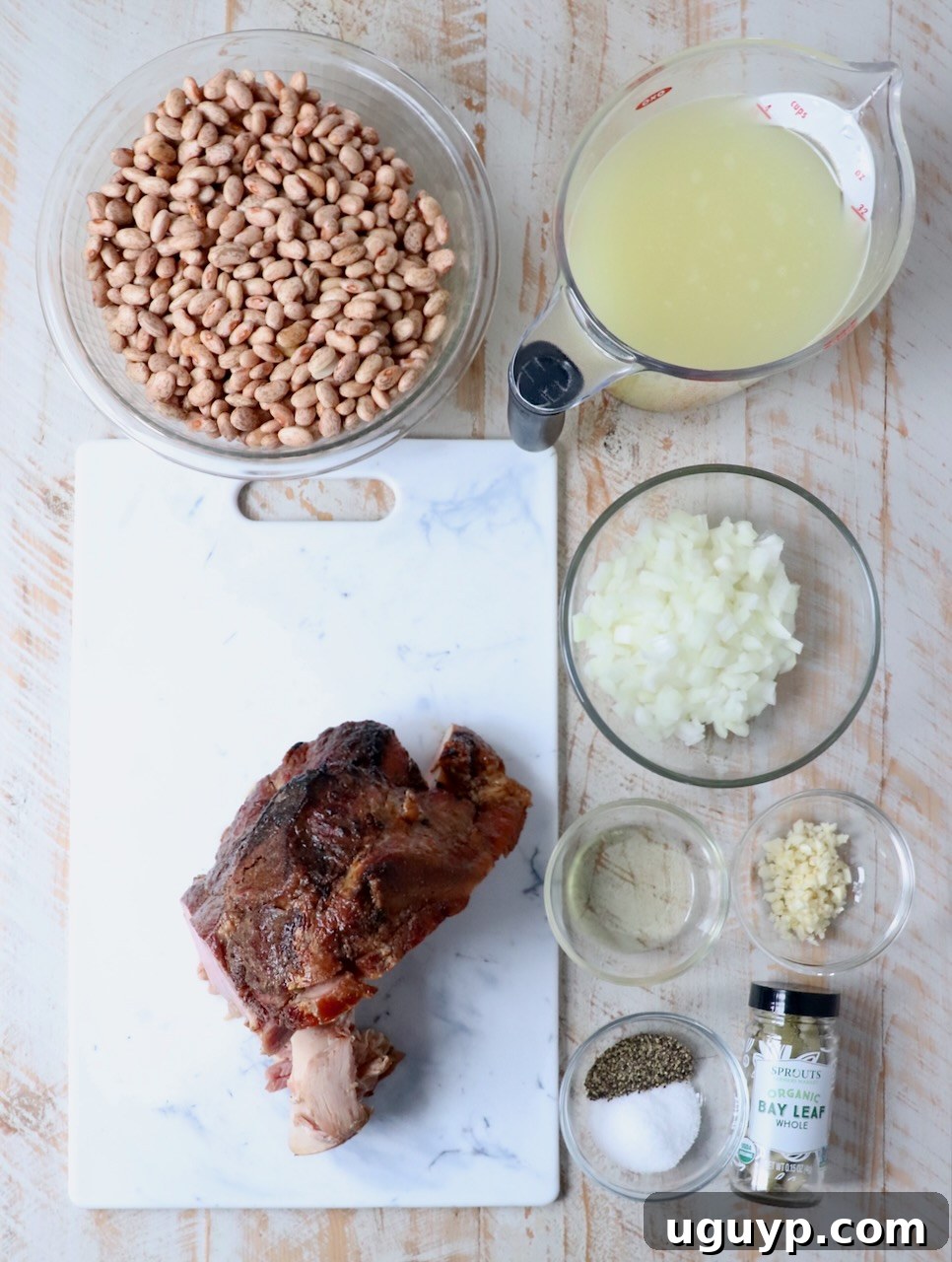 Assorted ingredients for Southern pinto beans laid out on a white wooden board, including dried pinto beans, onion, garlic, ham hock, and spices.