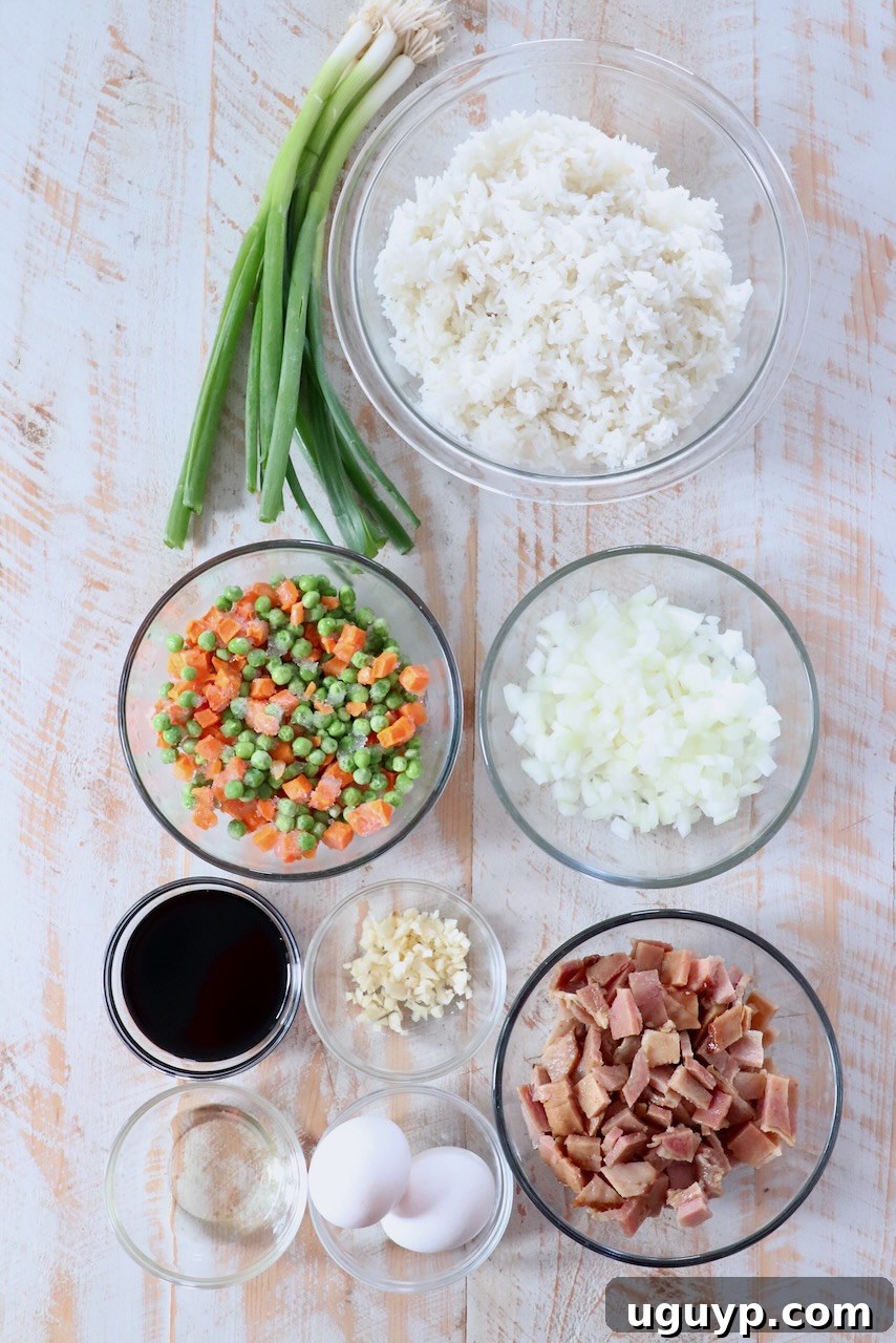 ingredients for ham fried rice on white wood board, including ham, rice, peas, carrots, onion, garlic, eggs, soy sauce, sriracha, and green onions