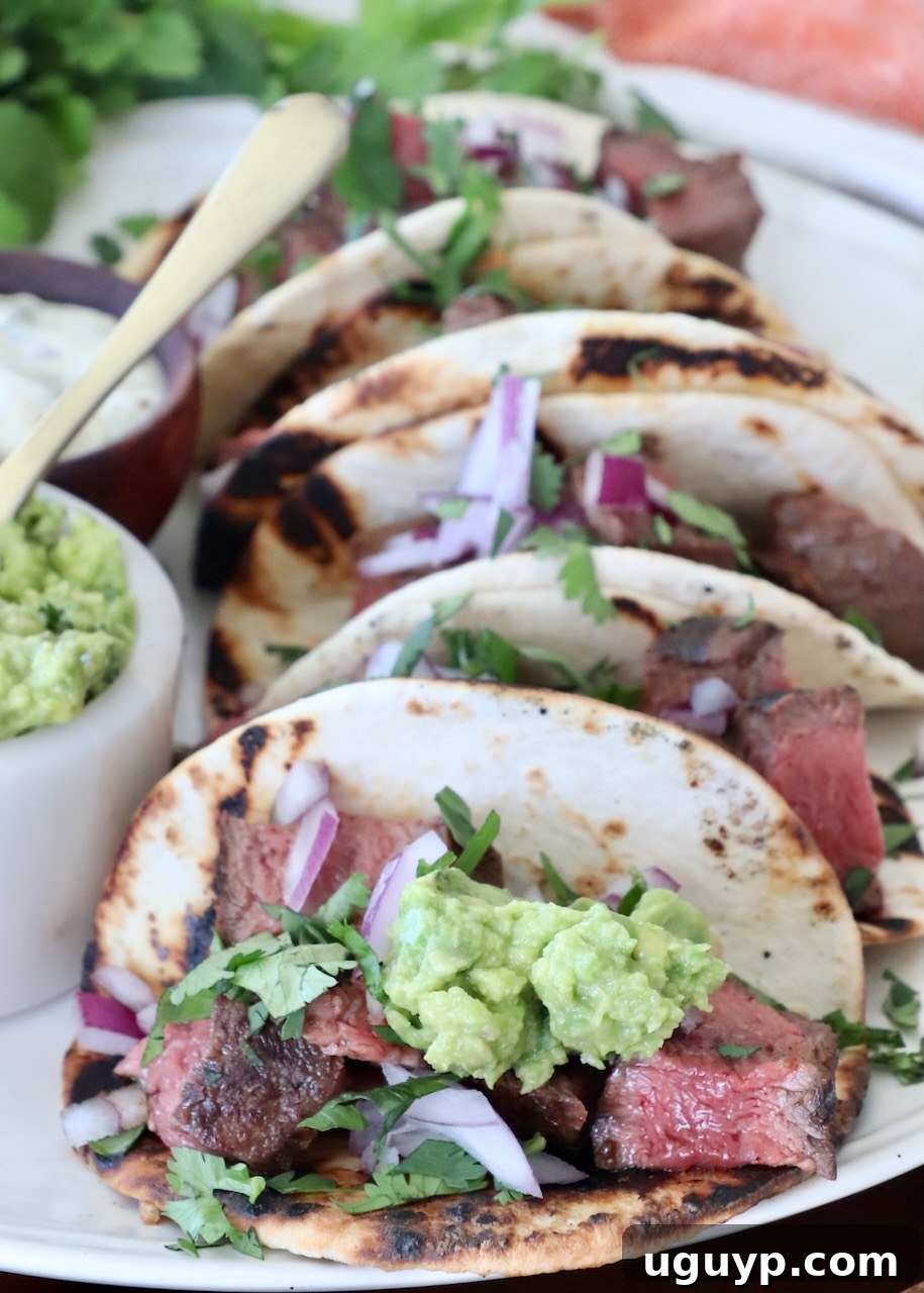 Two flat iron steak tacos on a plate, garnished with fresh cilantro and a dollop of guacamole, with a lime wedge.