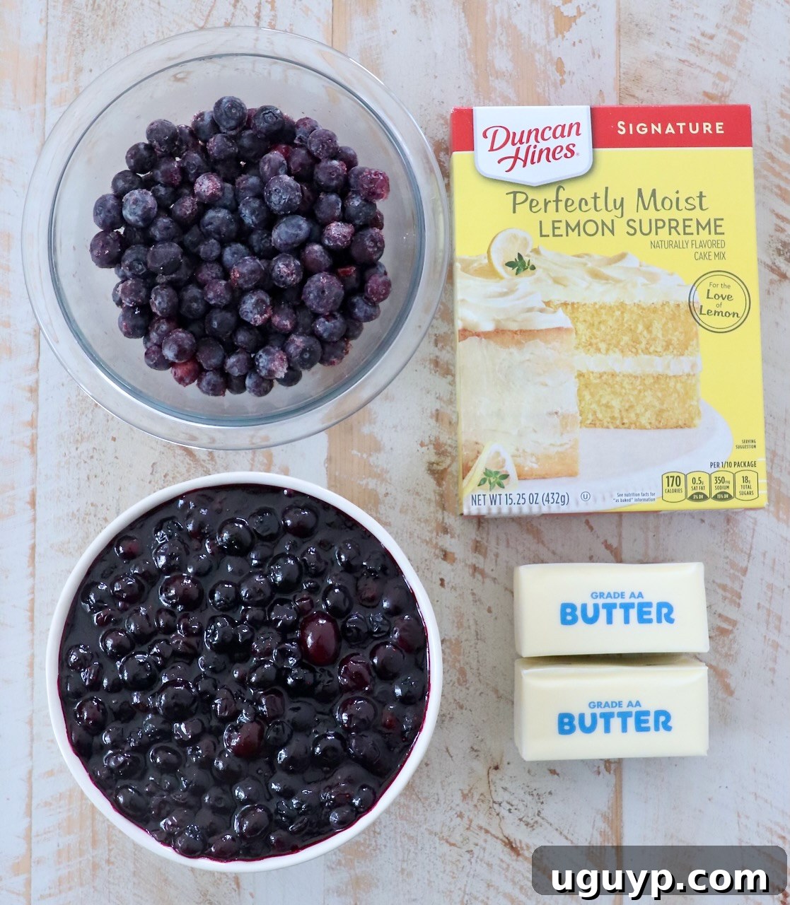 Four essential ingredients for blueberry dump cake: a can of blueberry pie filling, a box of lemon cake mix, a stick of butter, and a bowl of fresh blueberries, arranged on a rustic white wooden board.
