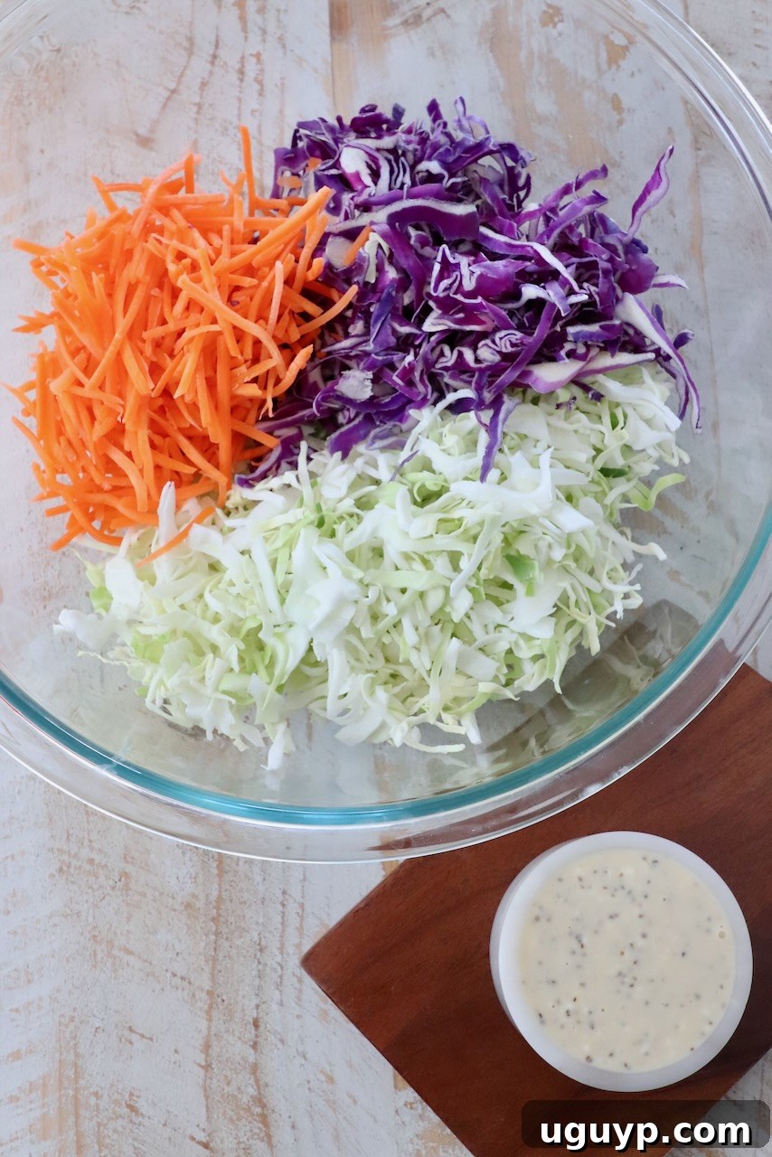 shredded cabbage and carrots in large glass bowl next to a small bowl of coleslaw dressing
