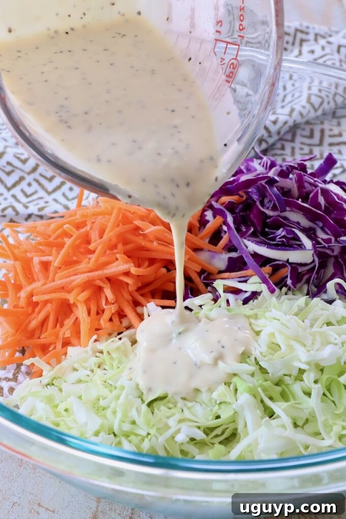 coleslaw dressing poured from glass measuring cup over cabbage in a large glass bowl