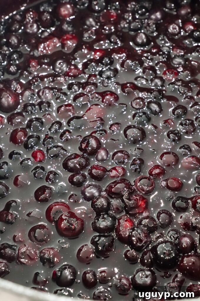 Blueberry pie filling simmering to a thick, dark consistency in a large saucepan, showing the cooked down blueberries.