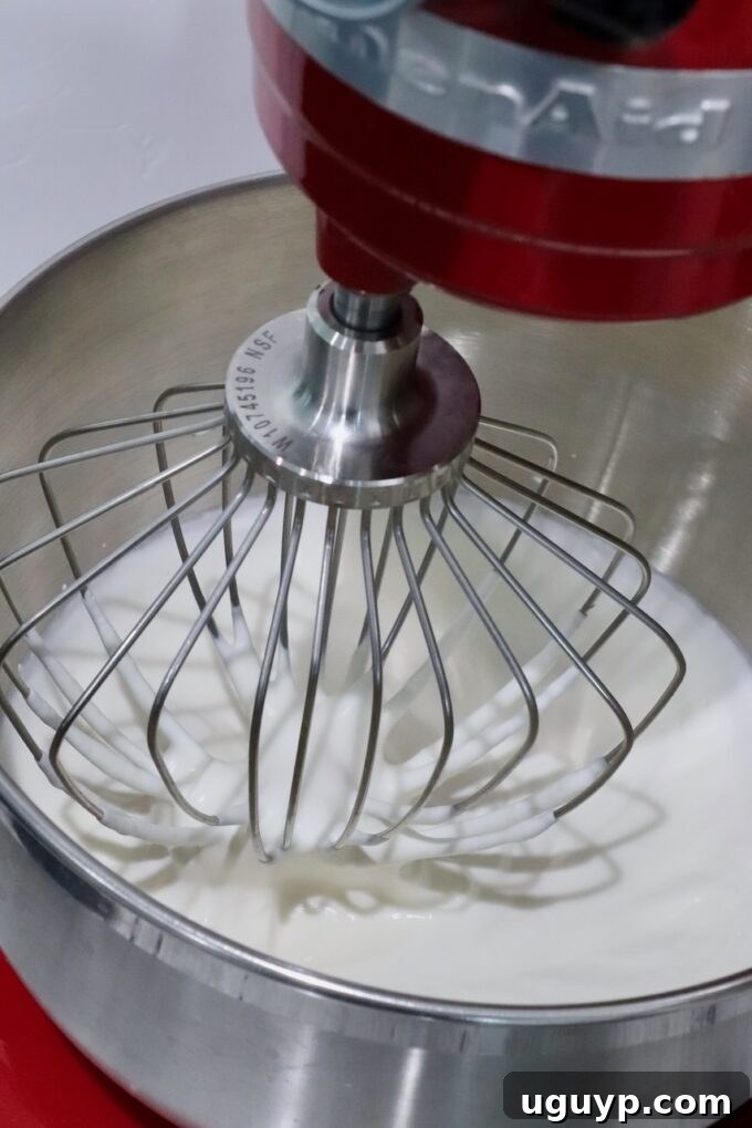 Cold heavy cream being whipped to firm peaks in a stand mixer with a whisk attachment.