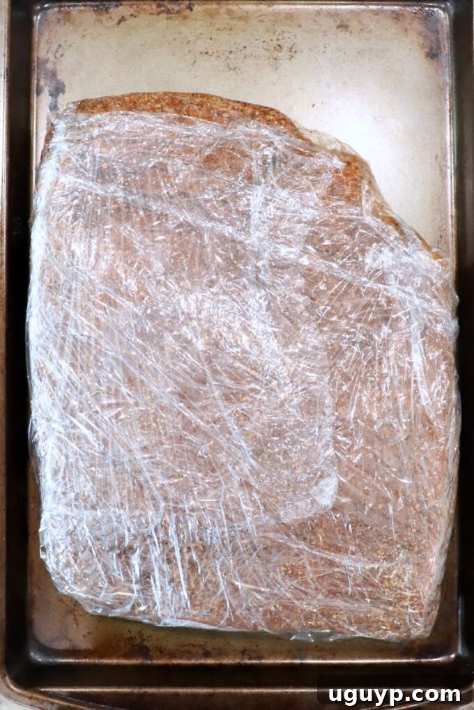 seasoned brisket wrapped tightly in plastic wrap on a baking sheet, ready for refrigeration