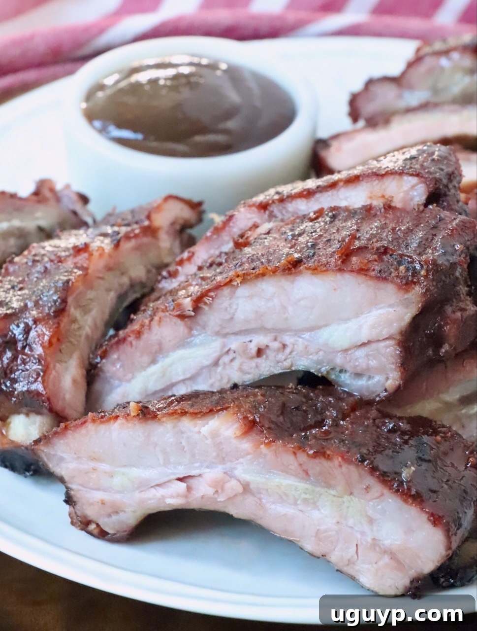 Sliced, smoky baby back ribs resting on a plate next to a small bowl of rich BBQ sauce, ready to be devoured.