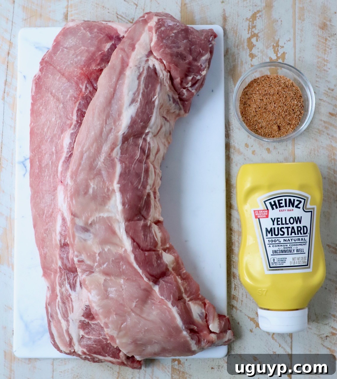 A vibrant display of ingredients for smoked ribs, including fresh baby back ribs, a bottle of apple juice, a bowl of dry rub, and a jar of mustard, arranged on a rustic white wooden board.