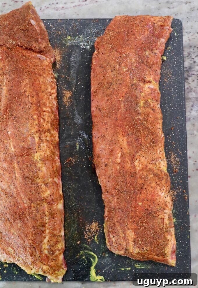 Two racks of baby back ribs generously coated with a savory dry rub, ready for the smoker, resting on a cutting board.