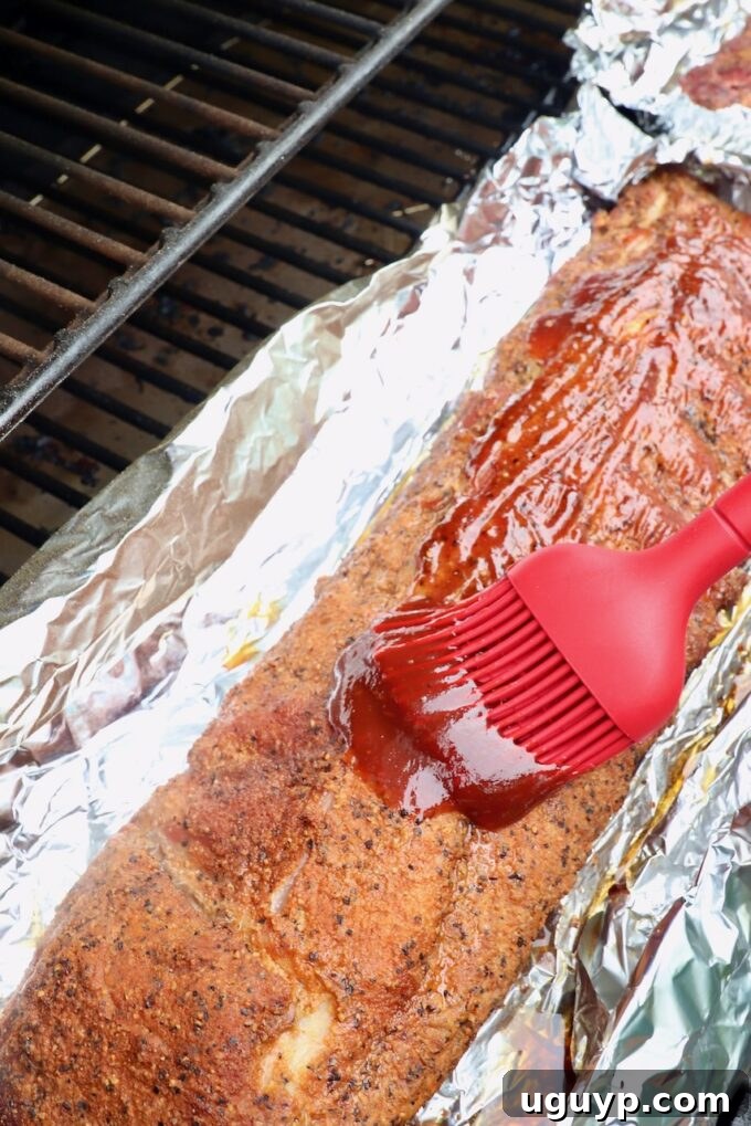 A kitchen brush expertly applying a generous layer of rich BBQ sauce to a rack of smoked ribs, still on foil, ready for the final glazing phase in the smoker.