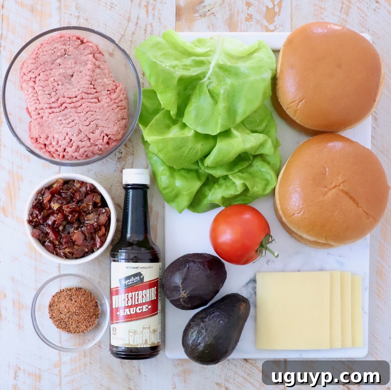 Ingredients laid out for California burgers on a pristine white wooden board, featuring ground beef, hamburger seasoning, Worcestershire sauce, white cheddar cheese, brioche buns, ripe avocados, fresh butter lettuce, juicy tomatoes, and a jar of homemade bacon jam.