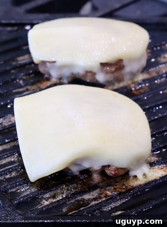 Burger patties topped with slices of white cheddar cheese, melting perfectly on the grill pan during the final minutes of cooking, creating a creamy, golden blanket over the beef.