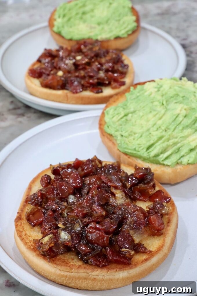 Toasted hamburger buns prepared on plates, with the bottom halves spread with rich bacon jam and the top halves smeared with creamy smashed avocado, ready for burger assembly.
