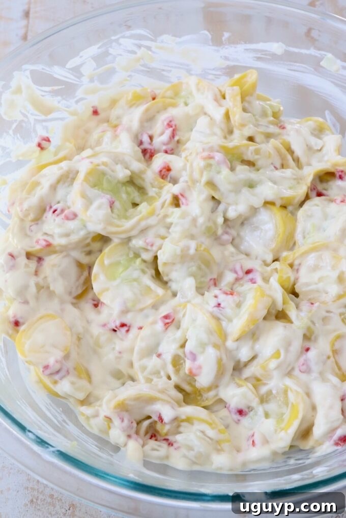 Drained, cooked yellow squash and onions stirred into the creamy mixture of cream of chicken soup, sour cream, and pimentos in a glass bowl.