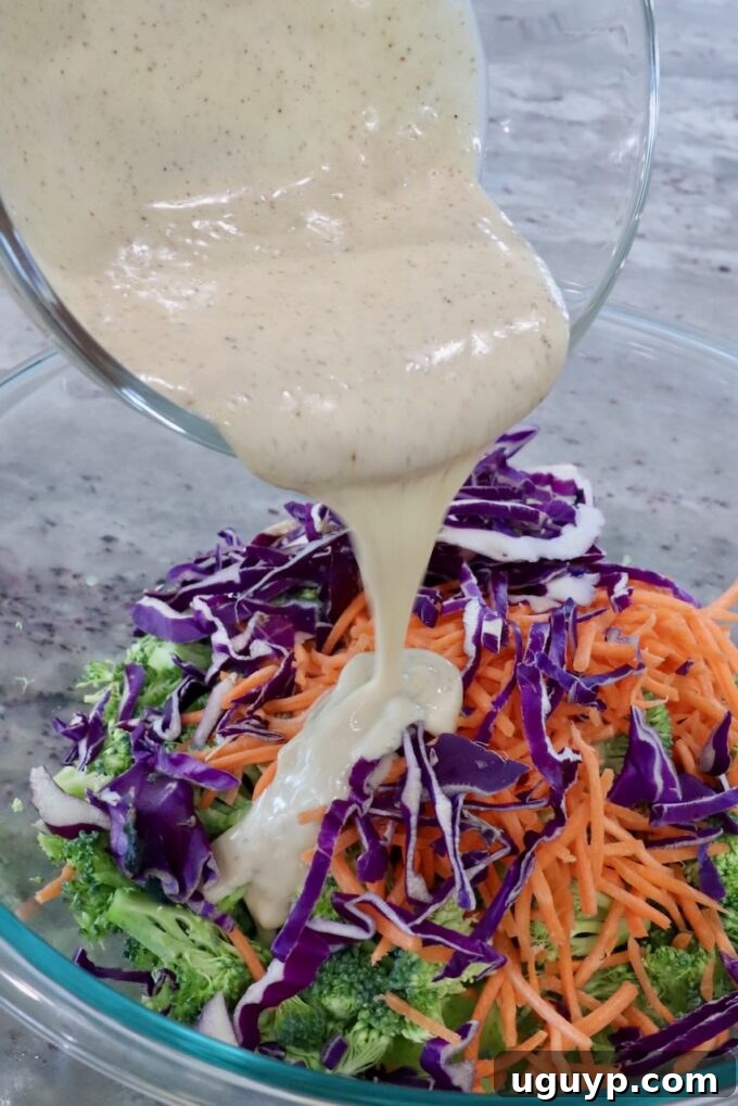Broccoli slaw in a bowl with the creamy dressing being poured over, ready for tossing.
