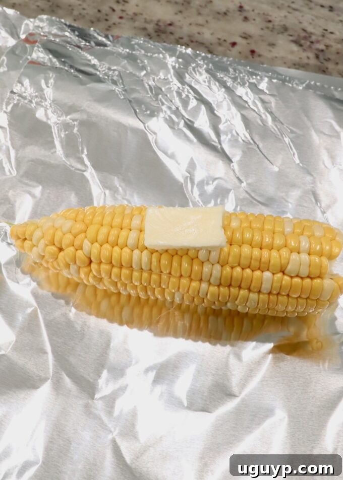 Shucked corn on the cob on aluminum foil with a pat of butter, ready for grilling.
