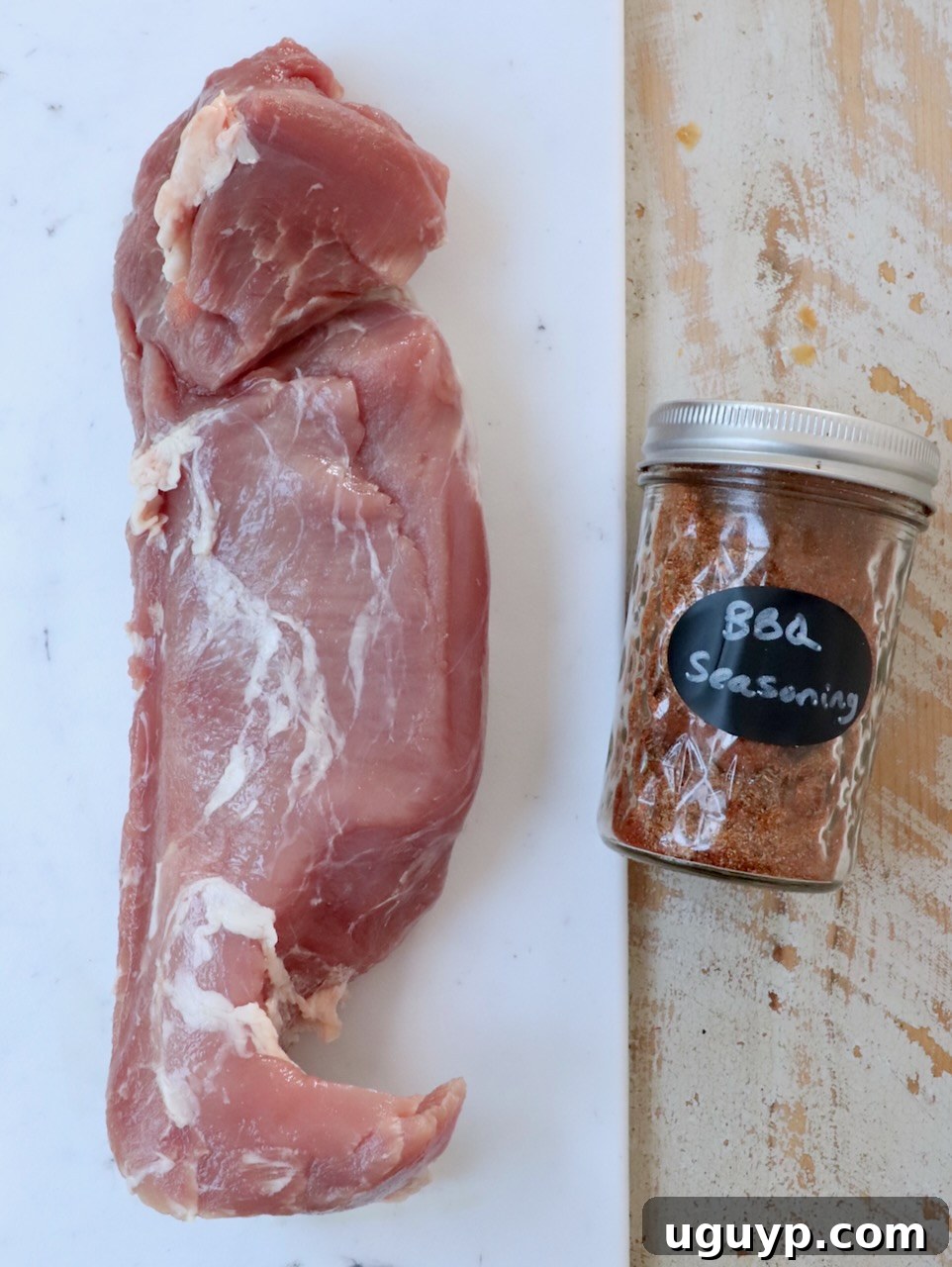 Raw pork tenderloin and BBQ seasoning on a white wood board.