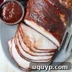 Sliced smoked pork loin on a plate with a side bowl of BBQ sauce, highlighting its tender texture and appealing color.