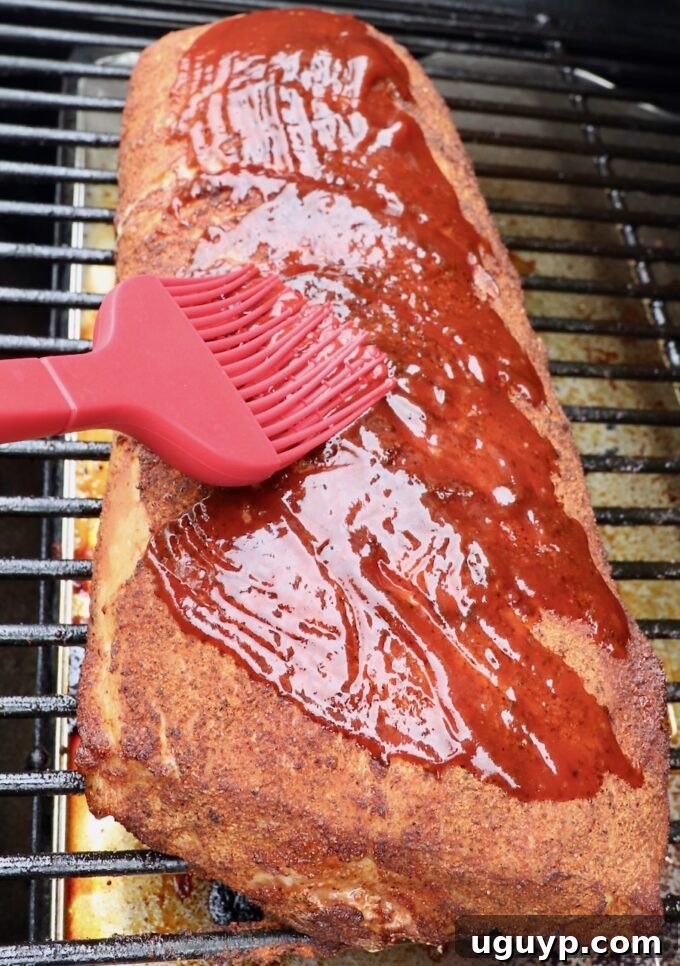 Pork loin on the smoker, with a basting brush applying a rich, flavorful layer of BBQ sauce as a finishing glaze, creating a beautiful caramelized exterior.