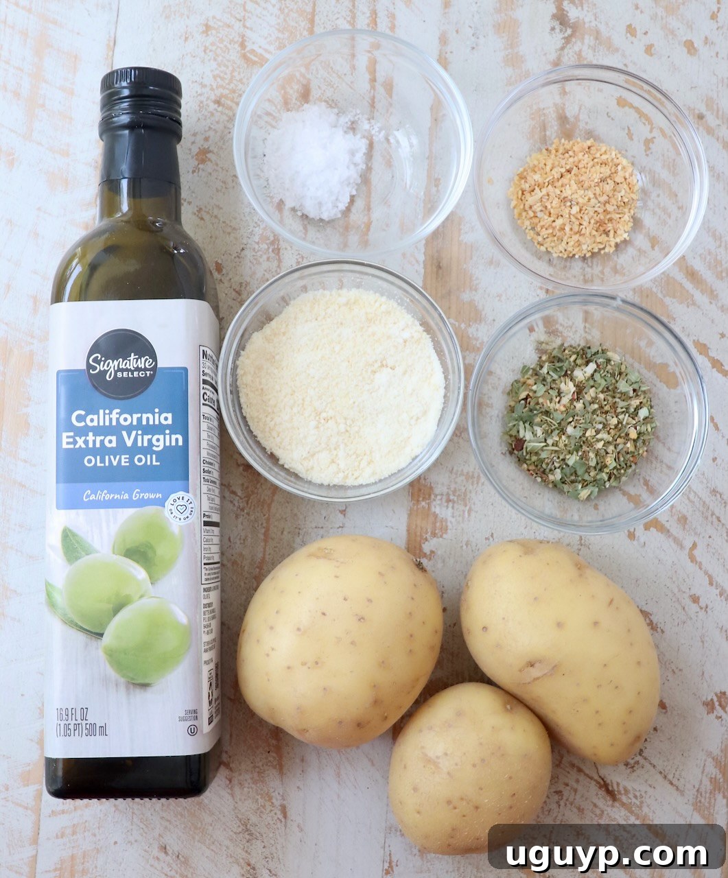 A collection of fresh ingredients for air fryer roasted potatoes, including Yukon gold potatoes, olive oil, Parmesan, Italian seasoning, sea salt, and dried minced garlic, neatly arranged on a white wooden board.