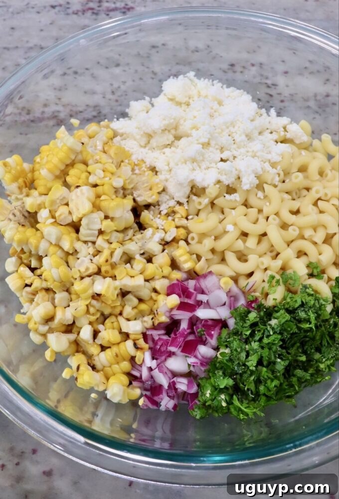 Cooked macaroni pasta in a large glass bowl with diced red onions, chopped cilantro, corn kernels, and crumbled cotija cheese before dressing.