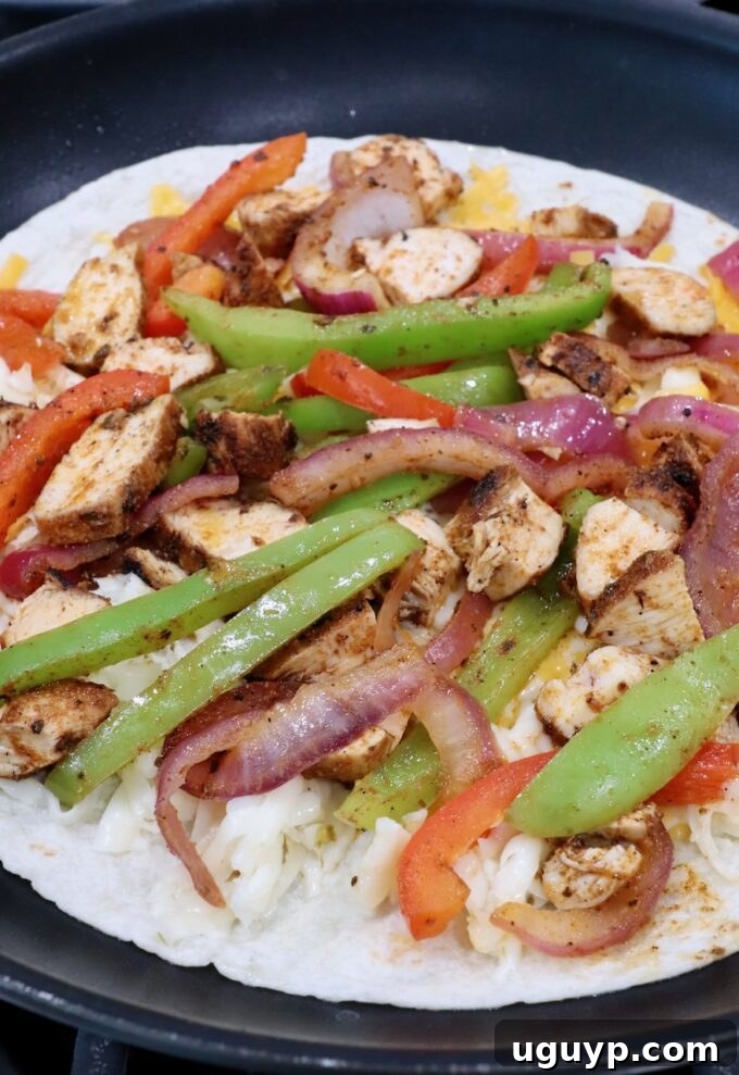 A tortilla in a skillet topped with a layer of cheese, followed by a savory mixture of sliced fajita vegetables and diced grilled chicken, perfectly arranged.