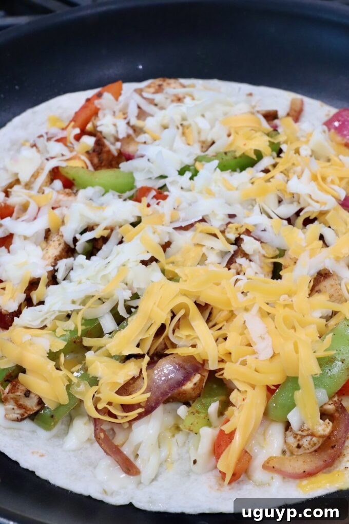 A fully loaded tortilla in a skillet, showcasing layers of cheese, fajita vegetables, diced chicken, and another layer of cheese, ready to be topped with a second tortilla.