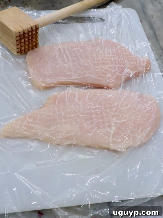 Chicken breasts on a cutting board covered with plastic wrap, with a meat tenderizer beside it.