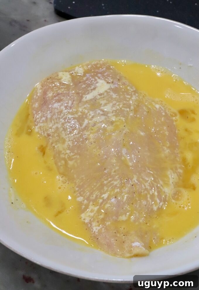 A chicken breast being dipped into a bowl of whisked eggs.