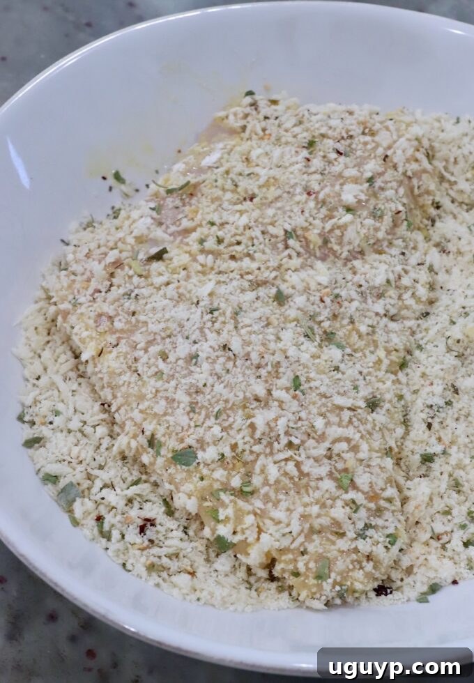 A chicken breast being pressed into a bowl of breadcrumbs mixed with Parmesan cheese and Italian seasoning.