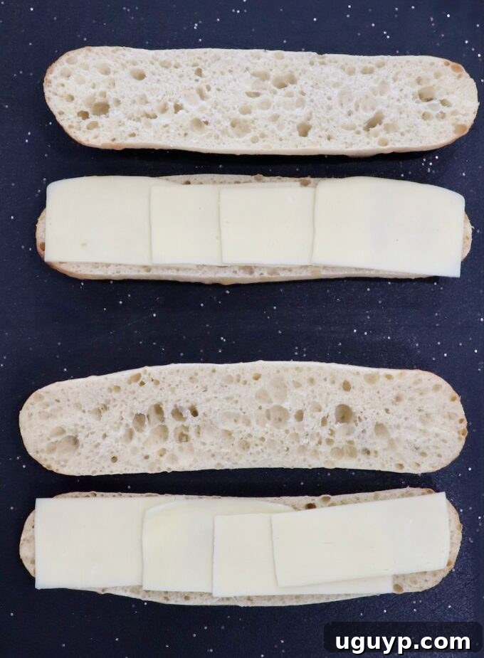 Two demi baguettes sliced open on a cutting board, topped with slices of mozzarella cheese.